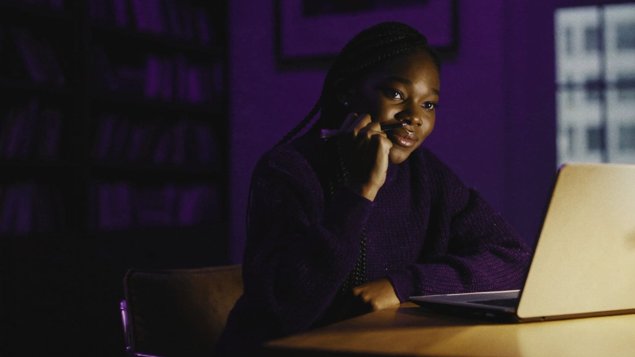 Une élève révise le soir devant son ordinateur portable, dans une bibliothèque baignée de lumière violette